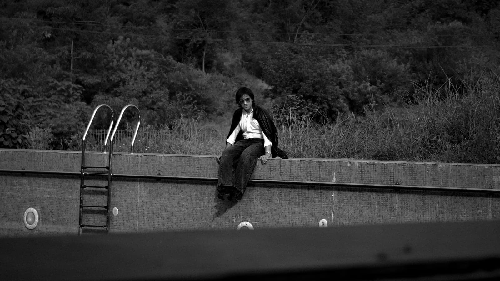 A lone figure sitting on a stone wall beside a ladder, in black and white